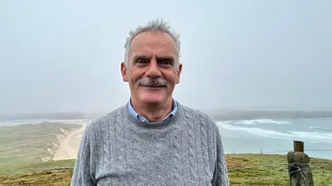 Andrew Ward, a man with short grey hair and a grey moustache, stands on high ground with a beach and stormy sea in the background. He is wearing a grey cable-knit sweater with a blue shirt underneath.