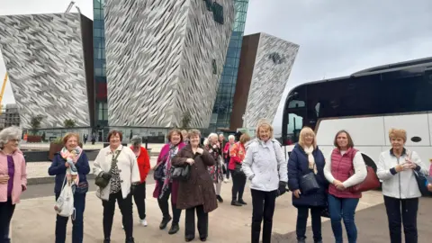 Lifestyle Development Group A group of women stand in front of the Titanic Belfast building. There are a<em></em>bout a dozen women wearing winter clothing. Behind them, beside Titanic Belfast, is a white coach.