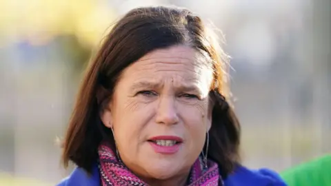 PA McDo<em></em>nald pictured with shoulder length brown hair. She stares at the camera as she wears pink lipstick, a pink scarf and a blue coat.